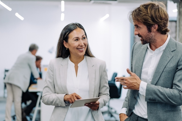 A smiling Asian female business leader holding a tablet, while walking with a white male colleague in a modern office with concrete pillars. Colleagues can be seen in the background