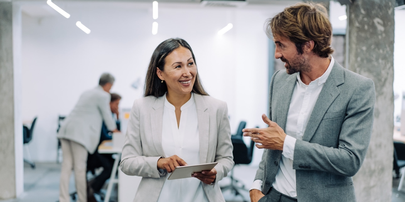 A smiling Asian female business leader holding a tablet, while walking with a white male colleague in a modern office with concrete pillars. Colleagues can be seen in the background