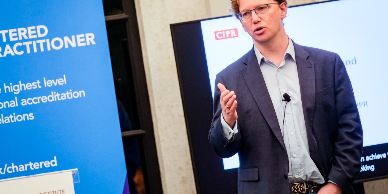 Steven Shepperson-Smith in a blue blazer addressing a CIPR conference. Behind him is a screen and to his left a blue display board