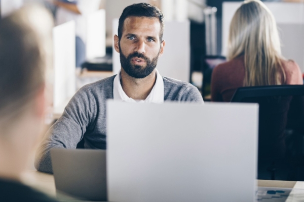 A white man with dark hair and beard sat at an office desk in conversation with a colleague whose back faces the camera.