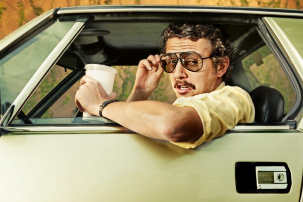 A white man with a moustache and curly hair raises his sunglasses as he leans out of a retro car while holding a coffee cup.