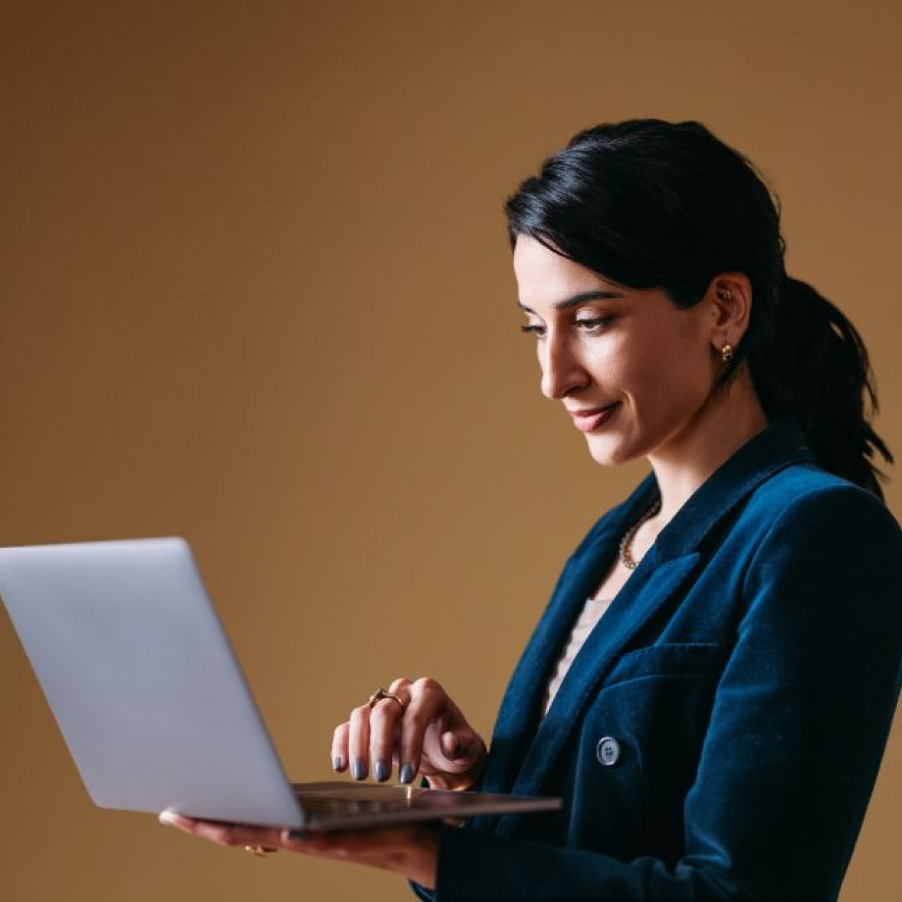 Against a brown background a business woman with dark hair and dressed in blue holds a laptop in her hand.