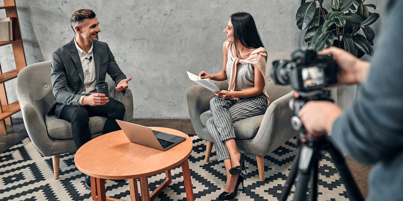 A white man in a grey suit holds a coffee cup and smiles while being interviewed by a white female journalist with long dark hair who wears a grey top and trousers. They are sat in armchairs with a table and laptop between them.