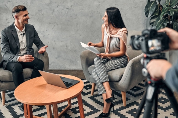 A white man in a grey suit holds a coffee cup and smiles while being interviewed by a white female journalist with long dark hair who wears a grey top and trousers. They are sat in armchairs with a table and laptop between them.