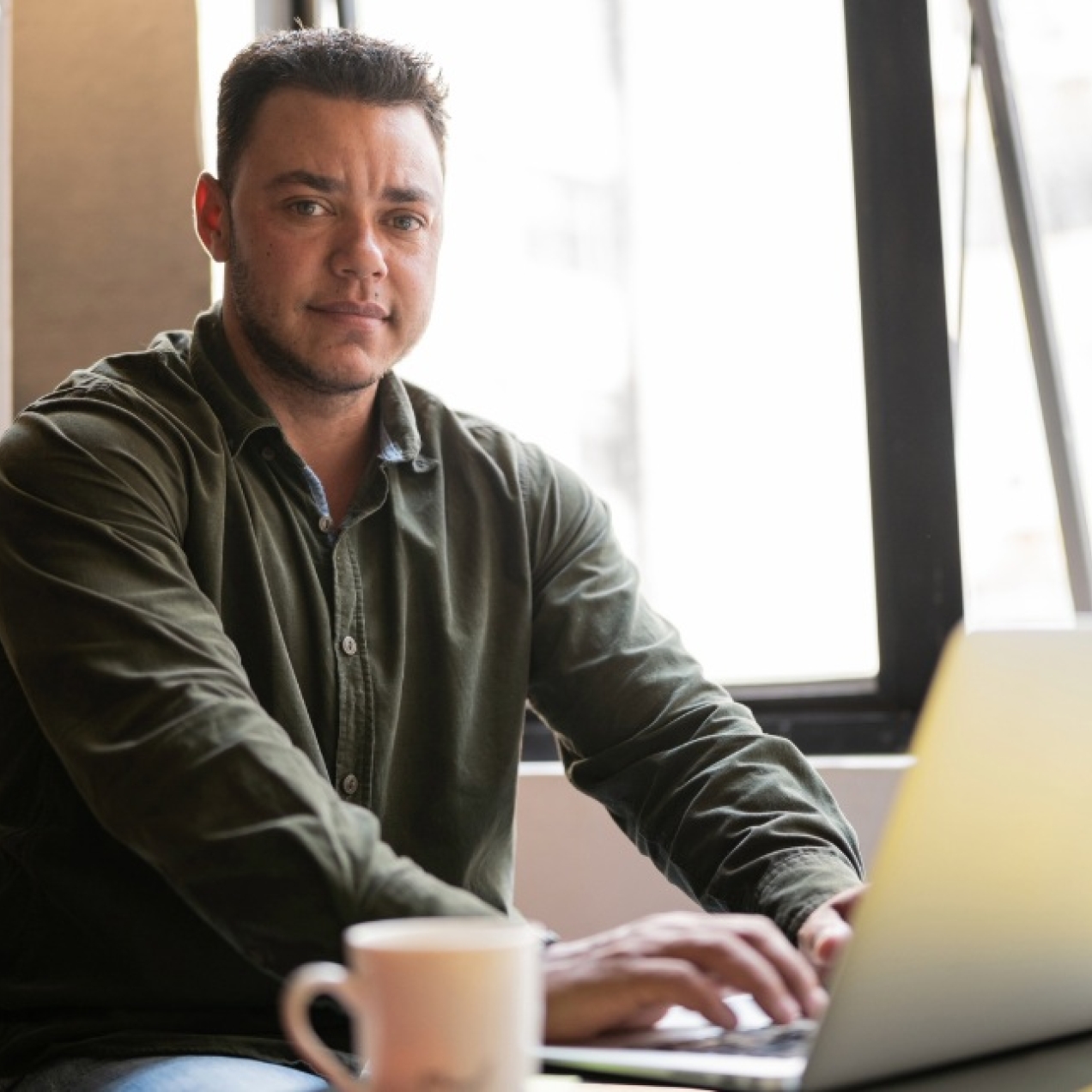 A transgender mature man with dark hair, who wears a dark green shirt, sat at a desk using a laptop in an office next to a window.