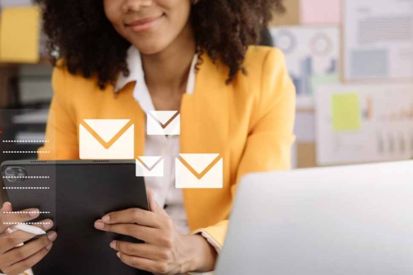 Envelope icons overlaid on a business woman wearing an orange jacket who is holding a tablet at a desk, with a laptop and clipboard beside her.