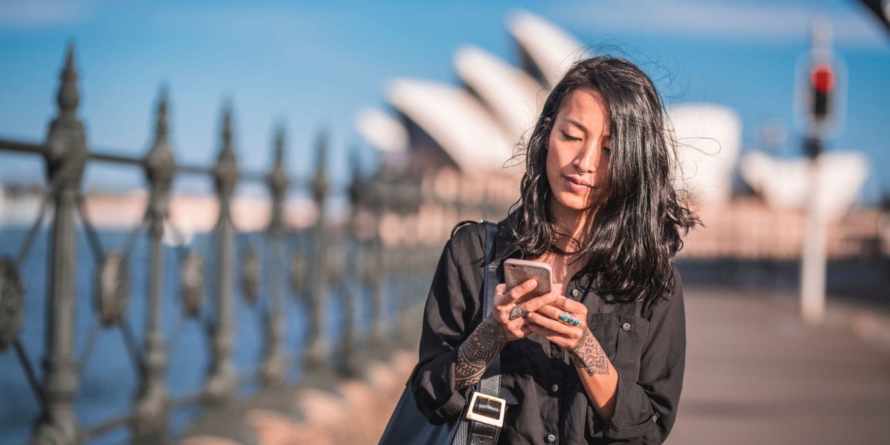 An Asian woman with long dark hair looks down at her mobile phone. The white sails of the Sydney Opera House are in the background against a blue sky.