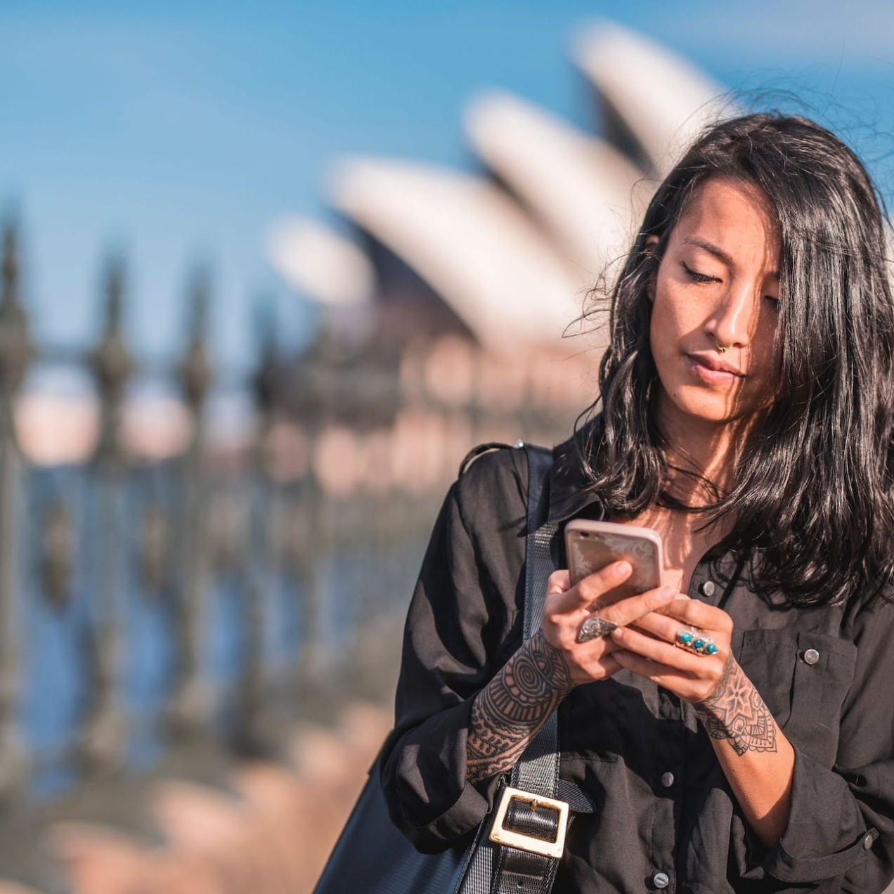 An Asian woman with long dark hair looks down at her mobile phone. The white sails of the Sydney Opera House are in the background against a blue sky.