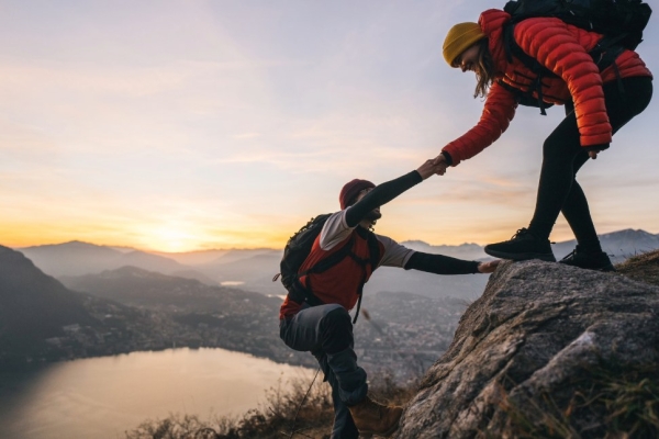 A female hiker offers a male hiker a hand up rocks on a mountain. A lake is visible in the distance below.