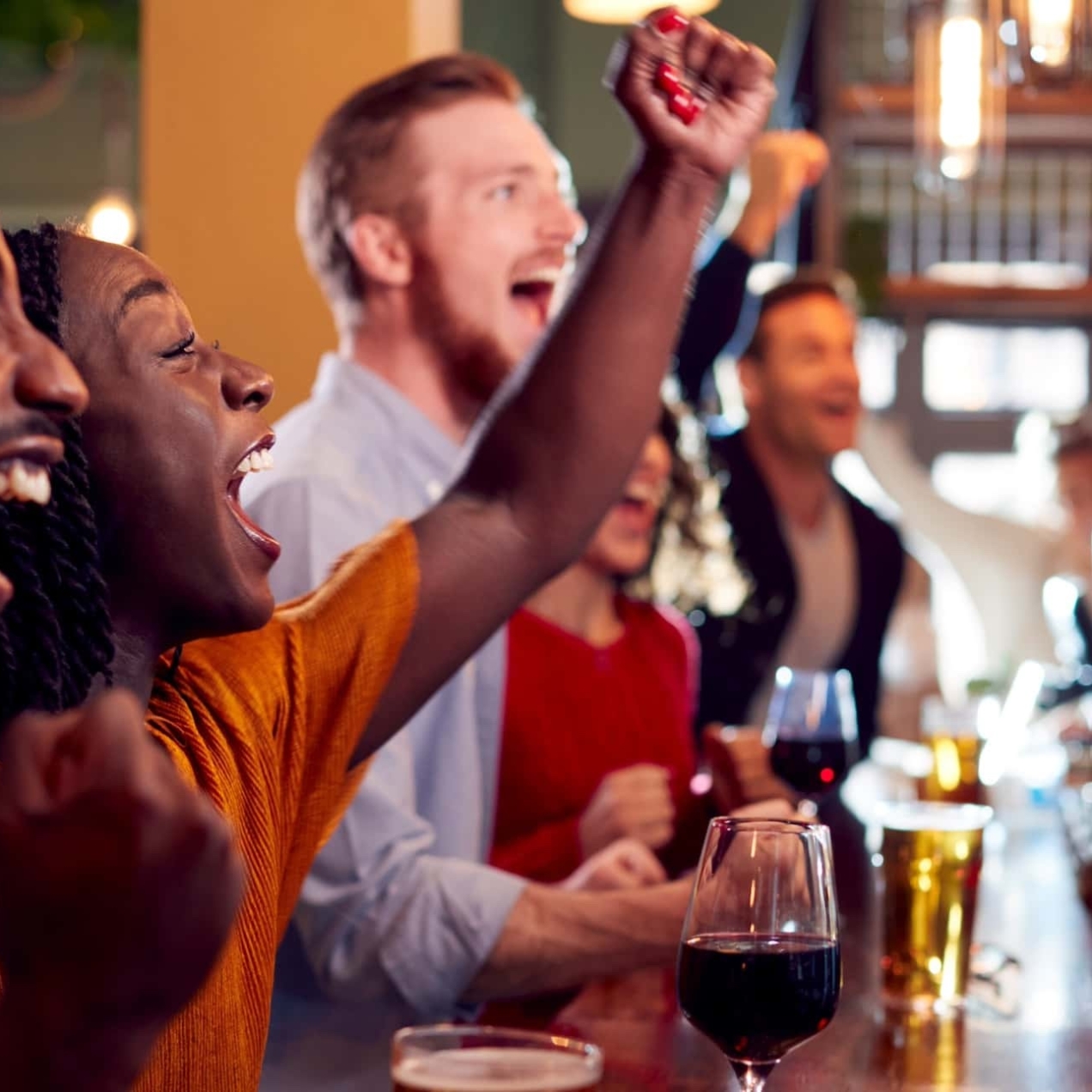 Stood at the bar of a pub, a Black man, Black woman two white men and a white woman clench their fists in a celebratory gesture while looking towards a screen which out of shot