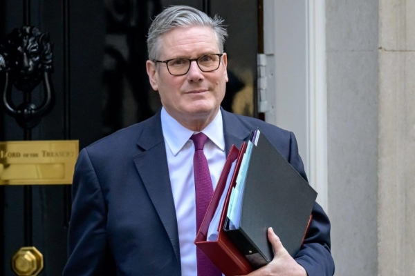 British prime minister Keir Starmer wearing a grey suit and holding a red folder. Behind him is the black door to 10 Downing Street.