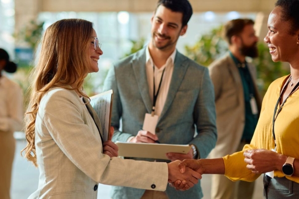 Two businesswomen shaking hands during a conference meeting, while a male colleague looks on.