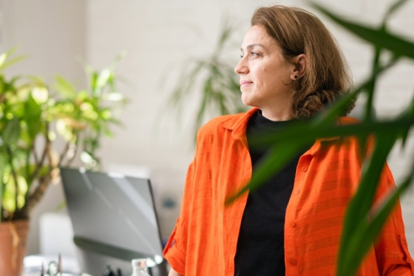 A middle-aged woman with brown hair and orange jacket over a black top looks to her right while stood in front of a desk in a light office. There are plants in the shot.