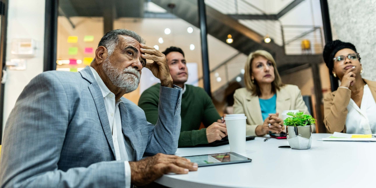 A worried Hispanic businessman with grey hair, beard and light blue suit, holds his hand to his head while concentrating on something out of shot. Colleagues - a man and a woman - sit next to him also looking pensive.