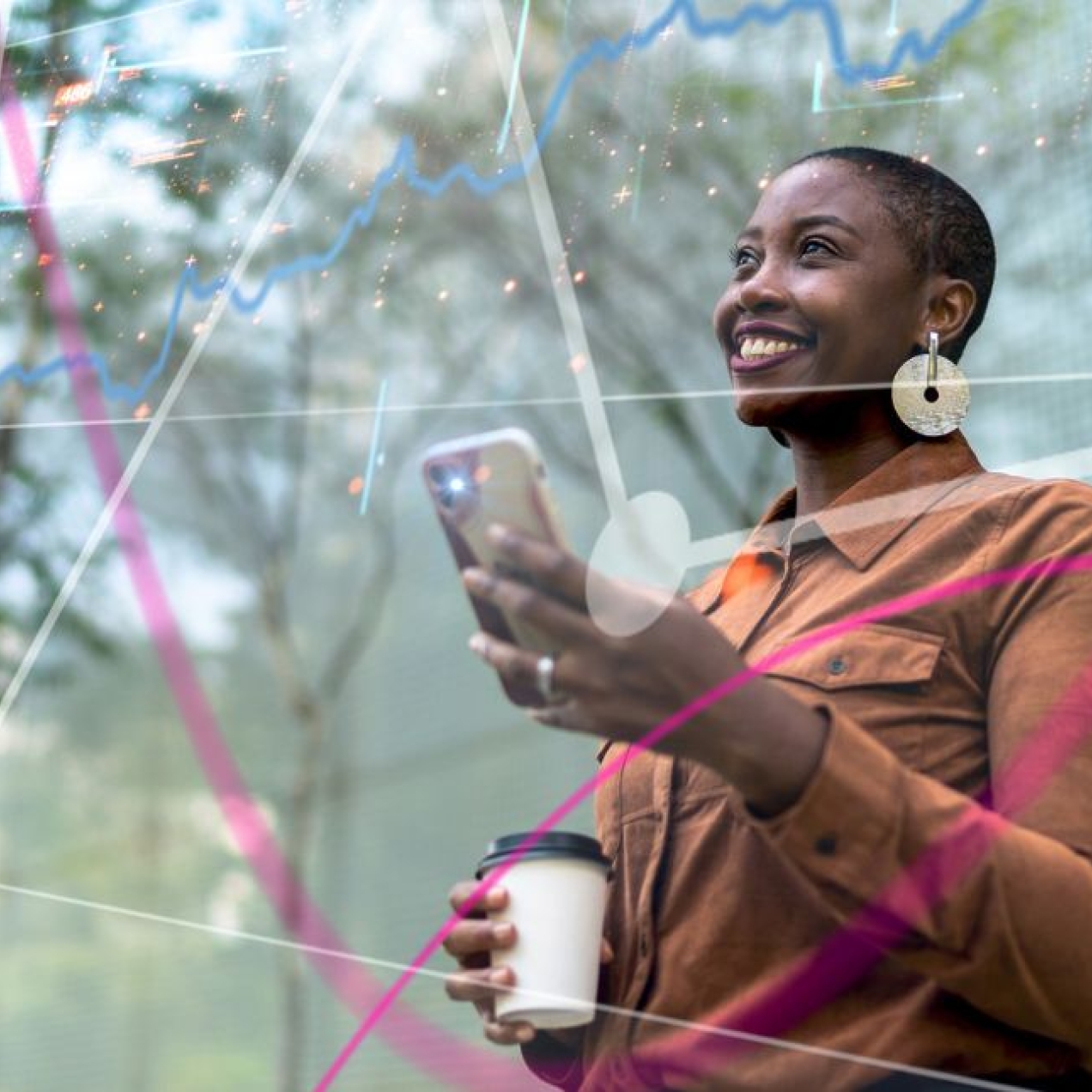 A smiling Black woman holds a phone in one hand and a coffee cup in the other, while stood beneath trees.