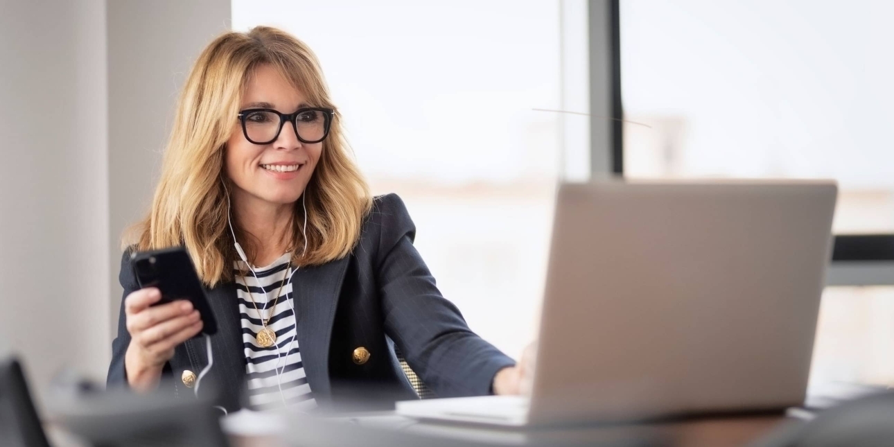 A middle-aged white woman with shoulder length blonde hair sits in front of a laptop with earphones connected to her mobile. She wears glasses and a navy jacket over a navy and white striped top