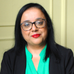 A colour portrait of Advita Patel against a cream background. Advita is an Asian woman with long dark hair who is wear glasses and a dark jacket over a mint-green shirt.