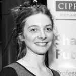 A black and white portrait of Amelia Stokes. Amelia is a white woman with dark hair who is smiling at the camera.