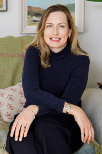 A colour portrait of Antonia Welch sat on a sofa with a green throw and picture behind her. Antonia is a white woman with long blonde hair wearing a black jumper and trousers.