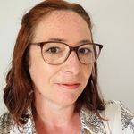 A colour portrait of Casey Freeman on a grey background. Casey is a white woman with shoulder length brown hair, who wears glasses.