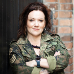 A colour portrait of Dannie-Lu Carr stood in front of a brick wall with her arms crossed. She is a white woman with dark hair and wears a green camouflage jacket