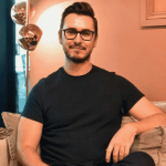A colour portrait of Dean Connelly sat in a pink room. Dean is a white man with dark hair and beard, who wears glasses and a dark shirt.