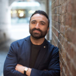 A colour portrait of Detch Singh stood with arms crossed in a brick alleyway. Hie is wearing a navy jacket.