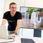 Dominic Ridley-Moy stood in front of a TV screen and laptop. Ridley-Moy is white man wearing a black t-shirt.