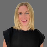 A colour portrait of Faith Hill on a grey background. Faith is a white woman with shoulder length blond hair and wearing a black top.