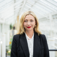 A colour portrait of Georgia Bailey, a white woman with long blonde hair, wearing a dark jacket over a white blouse. She is stood on a balcony under a glass roof.
