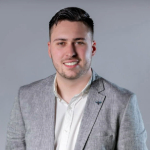 A colour portrait of Jack Richards against a grey background. Jack is a white man with short dark hair, wearing a grey blazer and a white shirt.