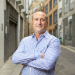 A colour portrait of Joshua Van Raalte in a narrow London street with his arms crossed. Joshua is a white man wearing a blue and white striped shirt.