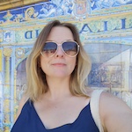 A colour portrait of Kat Dibbits wearing sunglasses. Kat is a white woman with shoulder length blonde hair who wears a blue top and is stood in front of blue and yellow ceramic tiles