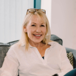 A colour portrait of Kate Everett sat on a sofa. Kate is a white woman with shoulder length blond hair, with glasses on the top of her head. She is wearing a white shirt.