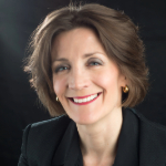 A head shot of Katherine Sykes against a black background. Katherine is a white woman with dark brown hair who is smiling and wearing a black jacket.