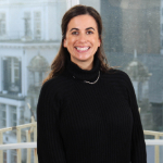 A colour portrait of Laura Rudolph smiling at the camera. Laura is a white woman with long dark hair who is wearing dark clothes.