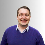 A colour portrait of Mario Creatura on a grey background. Mario is a white man white short brown hair wearing a purple jumper over a shirt.