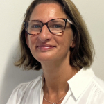 Miranda Smith smiles at the camera.She is a white woman with shoulder length light brown hair wearing glasses and a white shirt