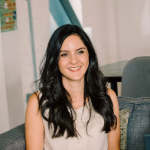 A colour portrait of Nikki Logan. Niki is a white woman with long dark hair, who is wearing a beige vest top.