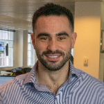 A colour portrait of Oli Foster, a white man with dark hair and stubble, wearing a blue and grey striped shirt.