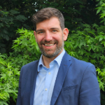 A colour portrait of Paul Compton. Paul is a white man with short brown hair, who wears a blue jacket over a light blue shirt. There are leaves in the background.