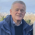 A colour photograph of Peter Osborne against a blue sky. Peter is a white man with grey hair wearing a blue coat and jumper.