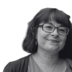 A black and white head shot of Rana Audah whois a white woman with dark hair wearing glasses, she is wearing a top and cardigan