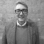 A black and white portrait of Richard Merrin against a brick wall. Richard is a white man with short hair and glasses who wears a blazer over a jumper.