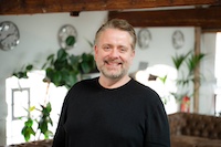 A colour portrait of Richard Stone, a white man with grey hair and beard, who wears a black jumper. In the background are plants and four clocks on a white wall