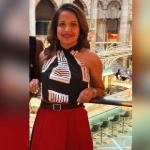 A colour portrait of Rishini Weeraratne stood in a restaurant. She wears a black patterned vest top and red skirt.