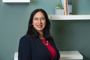 Sarah Liddiard is stood smiling in front of white shelves on an aquamarine wall. Sarah has long dark hair and wears a red top under a dark jacket