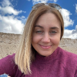 A colour portrait of Sarah Ion. Sarah is a white woman with blonde hair, wearing a blackcurrant jumper. There sky behind her is blue with white clouds.