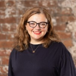 A colour photograph of Sarah Pinch stood in front of a brick wall. Sarah is a white woman with shoulder-length brown hair who wears glasses and a black top.