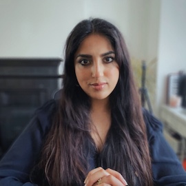 A portrait of Serena Hussain. Serena is an Asian woman with long dark hair, wearing a navy top. There is a black fireplace and white wall in the background